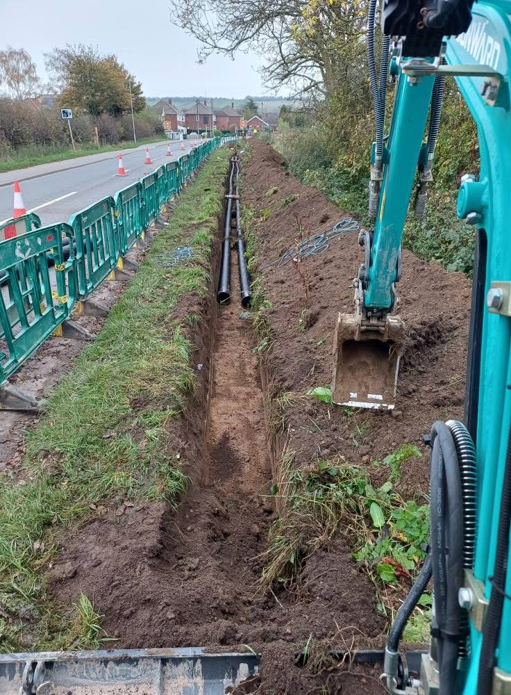 Excavated roadside utility trench with black ducting and a mini excavator